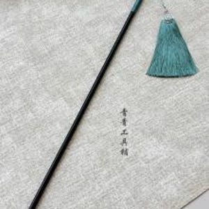 Pode incluir: Uma haste preta longa e fina com uma ponta verde-azulada e uma borla de tecido a condizer presa por um cordão fino. A haste apresenta caracteres chineses verticais em tinta preta, 青青工具精, posicionados ao longo da parte inferior.