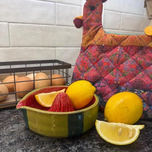May include: A ceramic citrus juicer with a green exterior and red interior, containing a whole lemon and a lemon wedge. Beside it sits a quilted fabric chicken-shaped tea cozy with a red, orange, and floral pattern, and a wire basket of eggs.