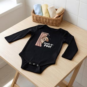 May include: A black long-sleeve baby bodysuit featuring a graphic of a light brown dog peeking from behind a pink shower curtain. The text "Peek-a-Poo!" is printed in white below the dog illustration. The garment has a snap closure at the bottom.