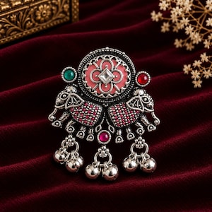 Könnte beinhalten: Ein Ring aus oxidiertem Silber mit zwei Elefantenfiguren mit rosa Steinakzenten, einem zentralen runden Medaillon mit rosa Emaille und einem klaren Stein, einem grünen Stein, einem roten Stein sowie drei hängenden silbernen Glöckchen.