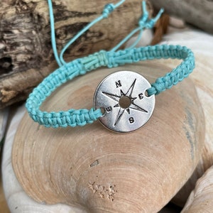 May include: A light blue macrame bracelet with a silver compass charm. The bracelet is displayed on a seashell. The compass charm has the cardinal directions marked. The bracelet is adjustable.