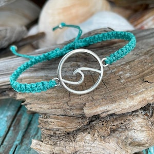 May include: A turquoise macrame bracelet with a silver wave charm. The bracelet is displayed on a piece of driftwood, with seashells in the background. The adjustable bracelet is approximately 7 inches in length.