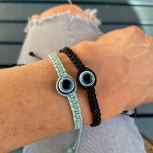 May include: Two macrame bracelets, one light blue and one black, each featuring a central evil eye charm. The bracelets are displayed on a person's arm, showcasing the intricate knotting and the detailed eye design.