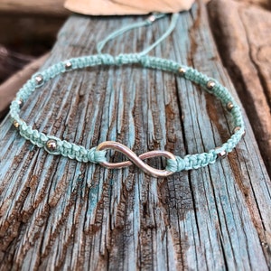 May include: A light blue macrame bracelet with a silver infinity symbol and small silver beads. The bracelet is displayed on a weathered, blue-painted wooden surface, with a seashell in the background. The bracelet is a handmade jewelry item.