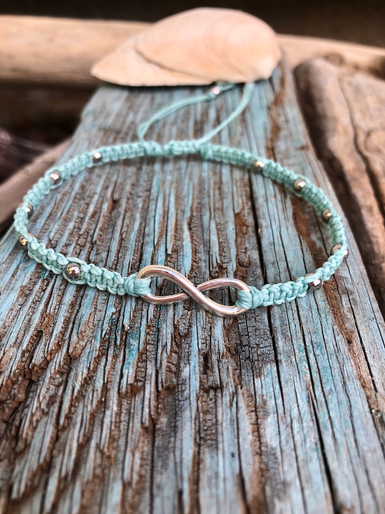 May include: A light blue macrame bracelet with a silver infinity symbol and small silver beads. The bracelet is displayed on a weathered, blue-painted wooden surface, with a seashell in the background. The bracelet is a handmade jewelry item.