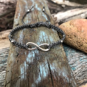 May include: A gray macrame bracelet featuring a silver infinity symbol. The bracelet has adjustable sliding knots and small silver beads. The bracelet is displayed on a weathered wooden surface with blue and gray tones.
