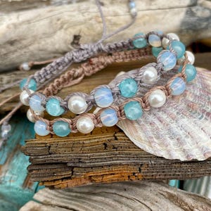 May include: Three macrame bracelets with beads in shades of blue, white, and opalescent. The bracelets are displayed on a piece of driftwood and a seashell. The bracelets have adjustable closures.