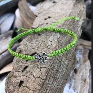 May include: A bright green woven bracelet with a silver four-leaf clover charm. The bracelet is made of a thick, braided cord and is displayed on a piece of weathered wood. The bracelet is adjustable.