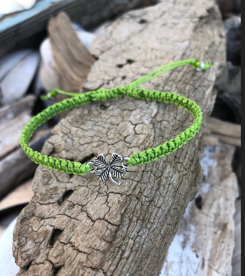May include: A bright green woven bracelet with a silver four-leaf clover charm. The bracelet is made of a thick, braided cord and is displayed on a piece of weathered wood. The bracelet is adjustable.