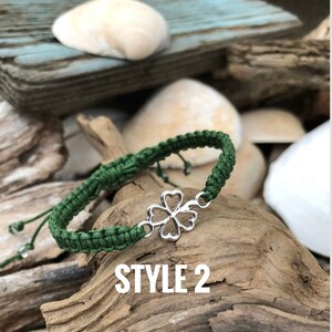 May include: A green macrame bracelet with a silver four-leaf clover charm. The bracelet is displayed on a piece of driftwood, with the text "STYLE 2" in white. The background includes seashells and weathered wood.