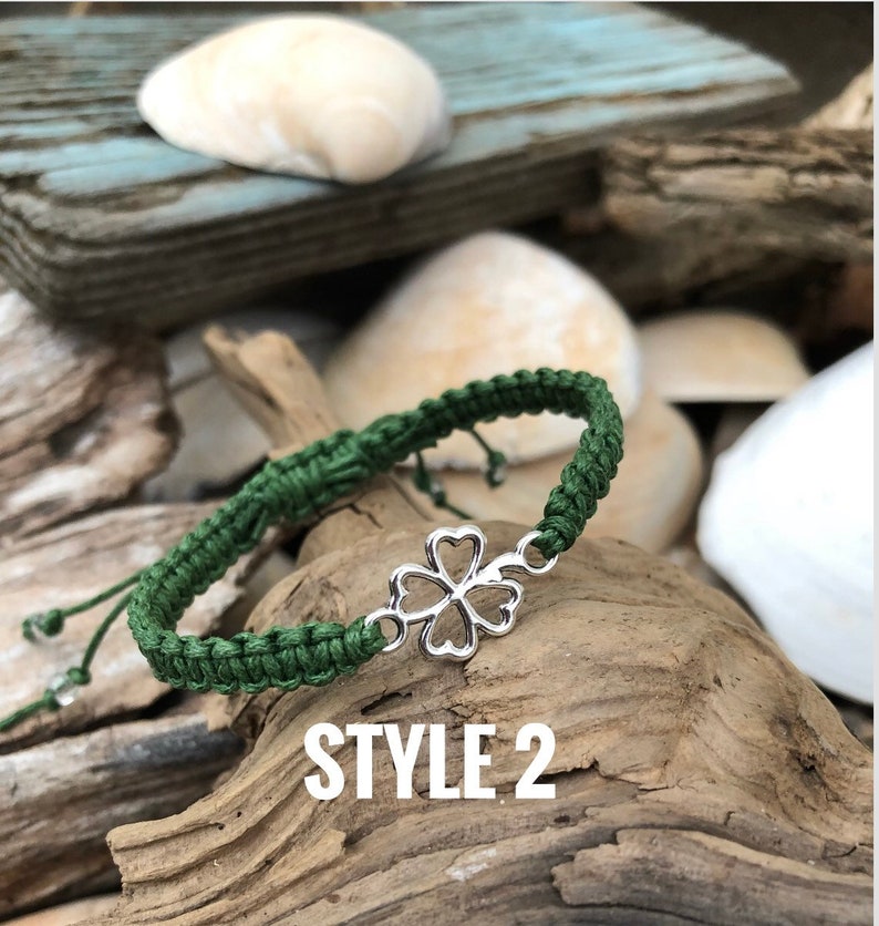 May include: A green macrame bracelet with a silver four-leaf clover charm. The bracelet is displayed on a piece of driftwood, with the text "STYLE 2" in white. The background includes seashells and weathered wood.
