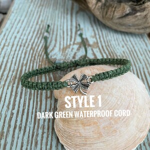 May include: Dark green waterproof cord bracelet with a silver-colored four-leaf clover charm. The bracelet is displayed on a seashell. Text on the image reads "STYLE 1 DARK GREEN WATERPROOF CORD."