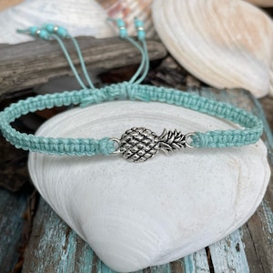 May include: A turquoise macrame bracelet with a silver pineapple charm. The adjustable bracelet features small turquoise beads at the ends. The bracelet is displayed on a white seashell, with a background of seashells and weathered wood.