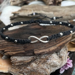 May include: Black macrame anklet featuring a silver infinity symbol and small silver beads. The adjustable anklet is displayed on a piece of driftwood. A stylish accessory for everyday wear.