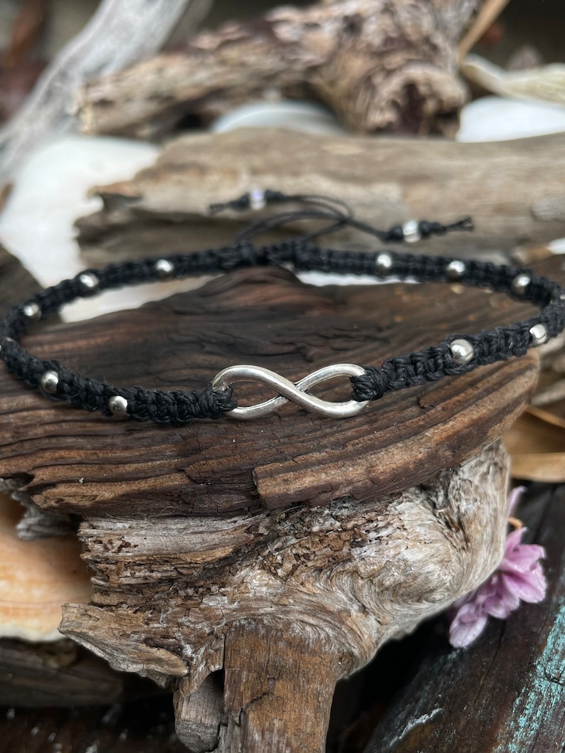 May include: Black macrame anklet featuring a silver infinity symbol and small silver beads. The adjustable anklet is displayed on a piece of driftwood. A stylish accessory for everyday wear.
