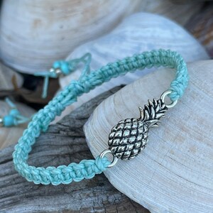 May include: A light blue macrame bracelet with a silver pineapple charm. The bracelet is displayed on a wooden surface with seashells in the background. The bracelet is adjustable.