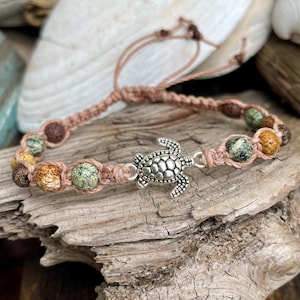 May include: A bracelet featuring a silver-toned metal sea turtle charm centered between a series of round, multi-colored stone beads. The beads display mottled patterns in shades of green, brown, and tan, held together by light brown cord.