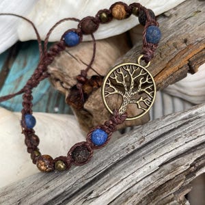 May include: Macrame bracelet featuring a bronze tree of life charm. The bracelet incorporates brown, blue, and tan beads, with a woven cord. The tree of life charm is a circular pendant with an intricate tree design.