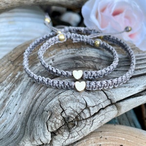 May include: Two gray macrame bracelets with gold heart charms and gold beads. The bracelets are displayed on a piece of driftwood, with a white rose in the background. The bracelets are adjustable.
