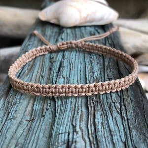 May include: A tan macrame bracelet with a woven design. The bracelet is displayed on a weathered, blue-painted wooden surface, with seashells in the background. The bracelet is adjustable.