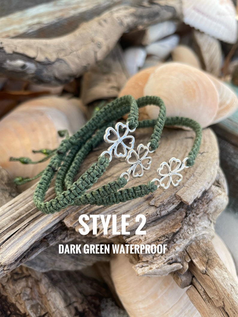 May include: Dark green waterproof bracelets with silver clover charms. The bracelets are made of woven cord and are displayed on driftwood with seashells in the background. The text "STYLE 2 DARK GREEN WATERPROOF" is visible.