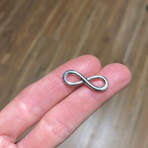 May include: A small, silver-colored infinity symbol. The figure eight shape is smooth and shiny, with rounded edges. The symbol is held between two fingers, with a wooden surface in the background.