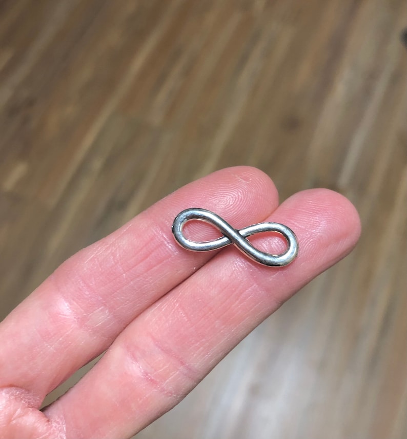 May include: A small, silver-colored infinity symbol. The figure eight shape is smooth and shiny, with rounded edges. The symbol is held between two fingers, with a wooden surface in the background.