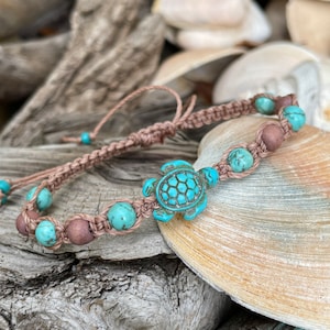 May include: Macrame bracelet featuring turquoise beads and a turquoise turtle charm. The bracelet also includes brown wooden beads. The adjustable closure is made of a light brown cord. This beach-themed bracelet is perfect for adding a touch of coastal style.