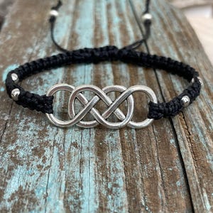 May include: Black macrame bracelet featuring a silver infinity knot charm. The bracelet has adjustable black cords and small silver beads. The infinity knot is a symbol of eternity and connection. This bracelet is a stylish accessory.