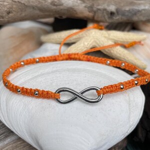 May include: Orange macrame bracelet featuring a silver infinity symbol. The bracelet is adorned with small silver beads. The bracelet is displayed on a white seashell, with driftwood and a starfish in the background.