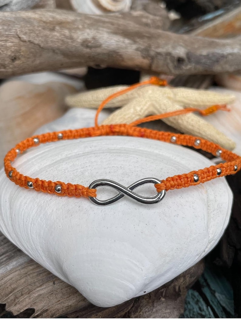 May include: Orange macrame bracelet featuring a silver infinity symbol. The bracelet is adorned with small silver beads. The bracelet is displayed on a white seashell, with driftwood and a starfish in the background.