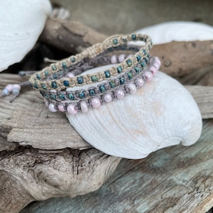 May include: Three handmade macrame bracelets with beads in shades of pink, blue, and gray. The bracelets are displayed on a seashell and weathered wood, creating a natural, beachy aesthetic. The bracelets are adjustable.