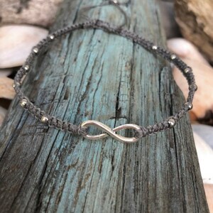 May include: A gray macrame anklet featuring a silver infinity symbol and small silver beads. The anklet is displayed on a weathered, blue-gray wooden surface. The adjustable design allows for a comfortable fit.