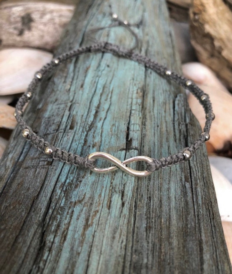 May include: A gray macrame anklet featuring a silver infinity symbol and small silver beads. The anklet is displayed on a weathered, blue-gray wooden surface. The adjustable design allows for a comfortable fit.