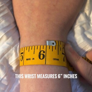 May include: A yellow measuring tape wrapped around a wrist, indicating a 6-inch circumference. The text "THIS WRIST MEASURES 6" INCHES" is displayed in white at the bottom of the image. The tape is marked with black numbers and lines.
