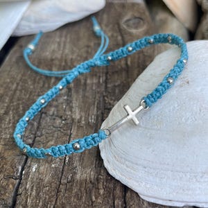 May include: A light blue macrame bracelet featuring a central silver-toned cross charm and small metallic beads woven throughout the cord. The bracelet includes adjustable pull strings at the ends, allowing for a flexible fit around the wrist.