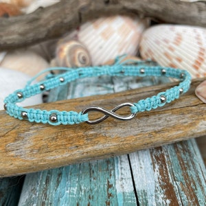 May include: A turquoise macrame bracelet with silver-colored beads and an infinity symbol charm. The bracelet is displayed on a piece of driftwood, with seashells in the background.