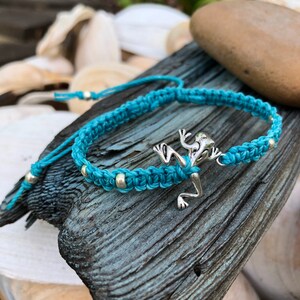 May include: A turquoise macrame bracelet with a silver frog charm and silver beads. The bracelet is displayed on a piece of weathered wood, with a blurred background of shells and greenery.