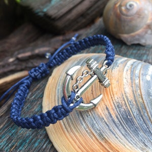 May include: A navy blue macrame bracelet with a silver anchor charm. The bracelet is knotted and features an adjustable closure. The anchor charm is detailed with a chain and is set against a seashell.