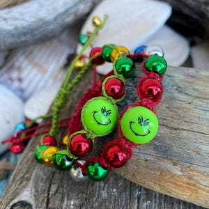 May include: Two handmade bracelets with red, green, silver, and gold beads. One bracelet features two large green beads with a smiling face. The bracelets are on a piece of driftwood with seashells in the background.