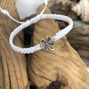 May include: White macrame bracelet with a silver four-leaf clover charm. The bracelet is adjustable and sits on a background of driftwood and seashells. A simple, handmade accessory.