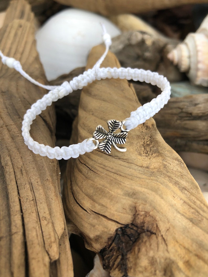 May include: White macrame bracelet with a silver four-leaf clover charm. The bracelet is adjustable and sits on a background of driftwood and seashells. A simple, handmade accessory.