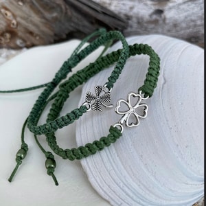 May include: Two green macrame bracelets with silver four-leaf clover charms. The bracelets are displayed on a white seashell. The macrame bracelets have adjustable sliding knots and are made of a dark green cord.