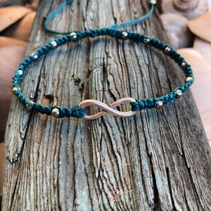 May include: Teal macrame bracelet featuring a silver infinity symbol. The bracelet is adorned with small silver beads and adjustable sliding knots. The jewelry is displayed on a piece of weathered wood, with seashells in the background.