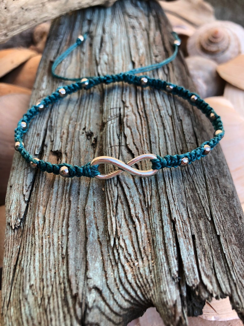 May include: Teal macrame bracelet featuring a silver infinity symbol. The bracelet is adorned with small silver beads and adjustable sliding knots. The jewelry is displayed on a piece of weathered wood, with seashells in the background.
