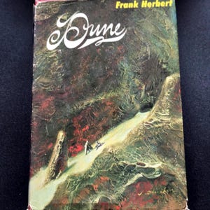 May include: A paperback book titled Dune by Frank Herbert with a textured, painterly cover illustration. The artwork features a winding light-colored path through a dark, abstract landscape of deep reds, browns, and greens with two small figures.