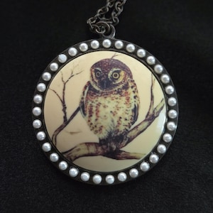 May include: A round pendant necklace featuring a cream-colored circular insert with a detailed illustration of a brown owl perched on a branch. The pendant is framed by a dark metal border accented with small white pearls and hangs on a chain.