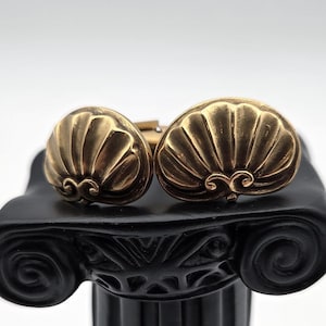 May include: A pair of brass-toned metal cufflinks featuring a sculpted shell design. Each cufflink has a semi-circular shape with vertical ridges radiating from a central decorative scroll element at the base, finished with a warm metallic sheen.