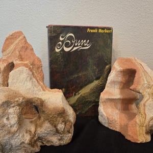 May include: A hardcover copy of the book Dune by Frank Herbert with a dark, illustrated cover, positioned between two large, porous, light brown and tan natural stone formations with visible layered textures and hollow openings.