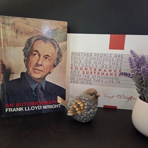 May include: A book titled An Autobiography by Frank Lloyd Wright, a white card with red accents featuring the text Whether people are fully conscious of this or not, they actually derive countenance and sustenance from the atmosphere of things they live in and with, a small stone-textured bird figurine, and a white pot with purple flowers.